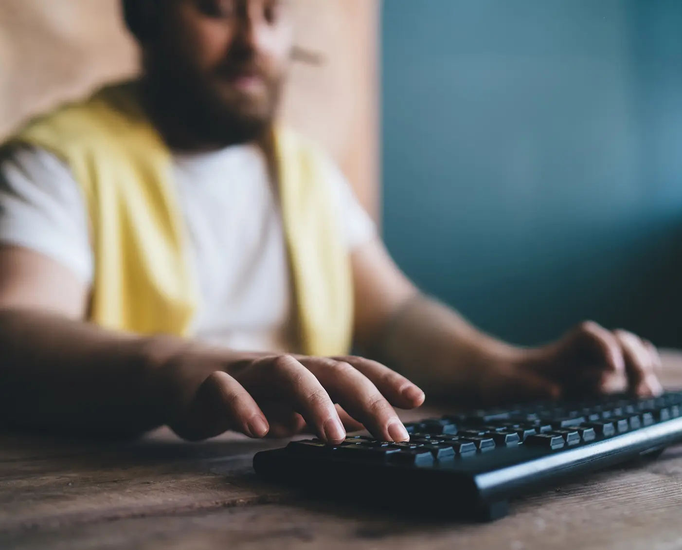 Close-up of hands typing on a black keyboard at a wooden desk. A yellow vest and white shirt are partially visible in the frame.