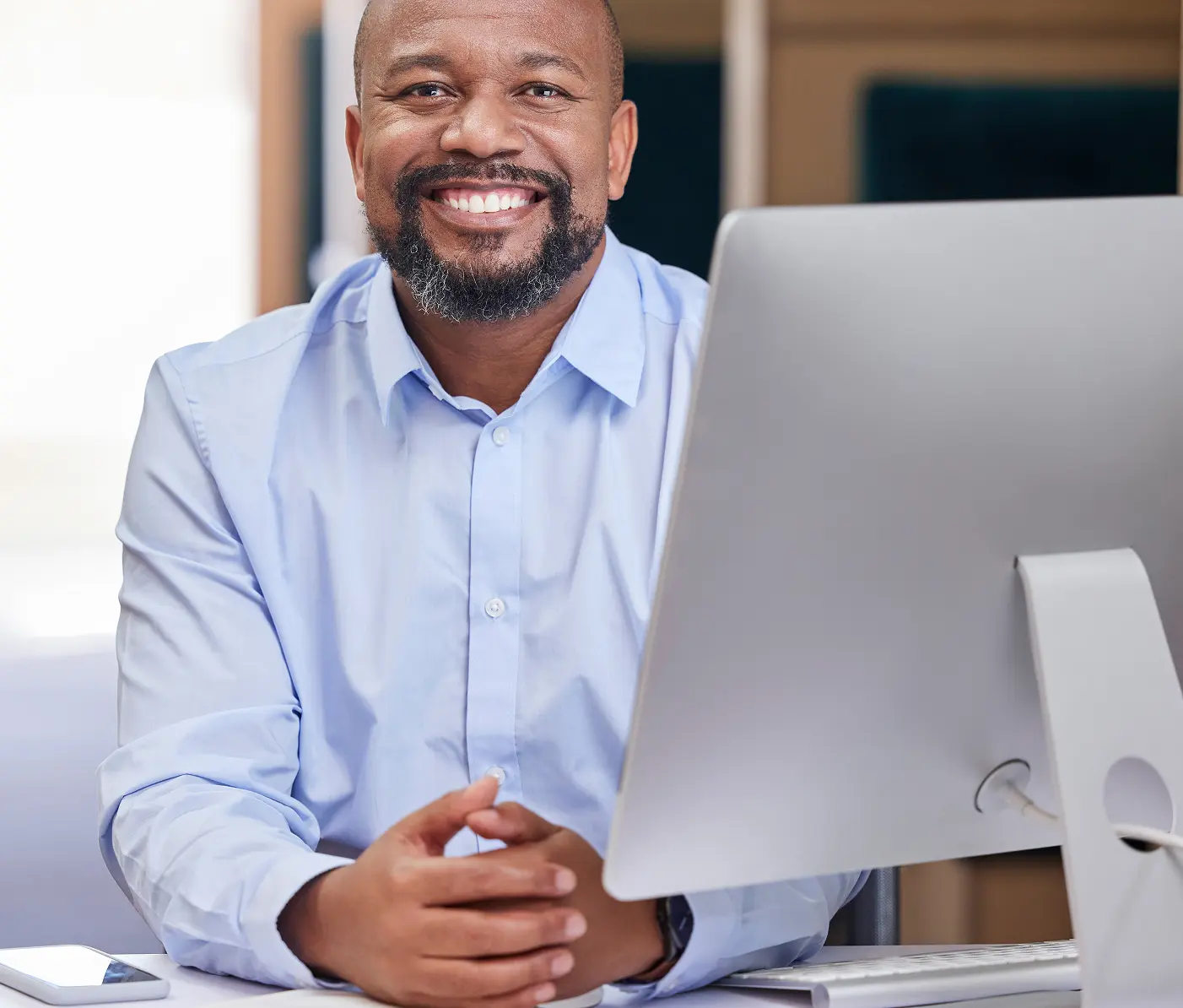 In a bright, well-lit office, a bearded man wearing a blue shirt is sitting at a desk with a computer monitor. He is smiling at the camera.