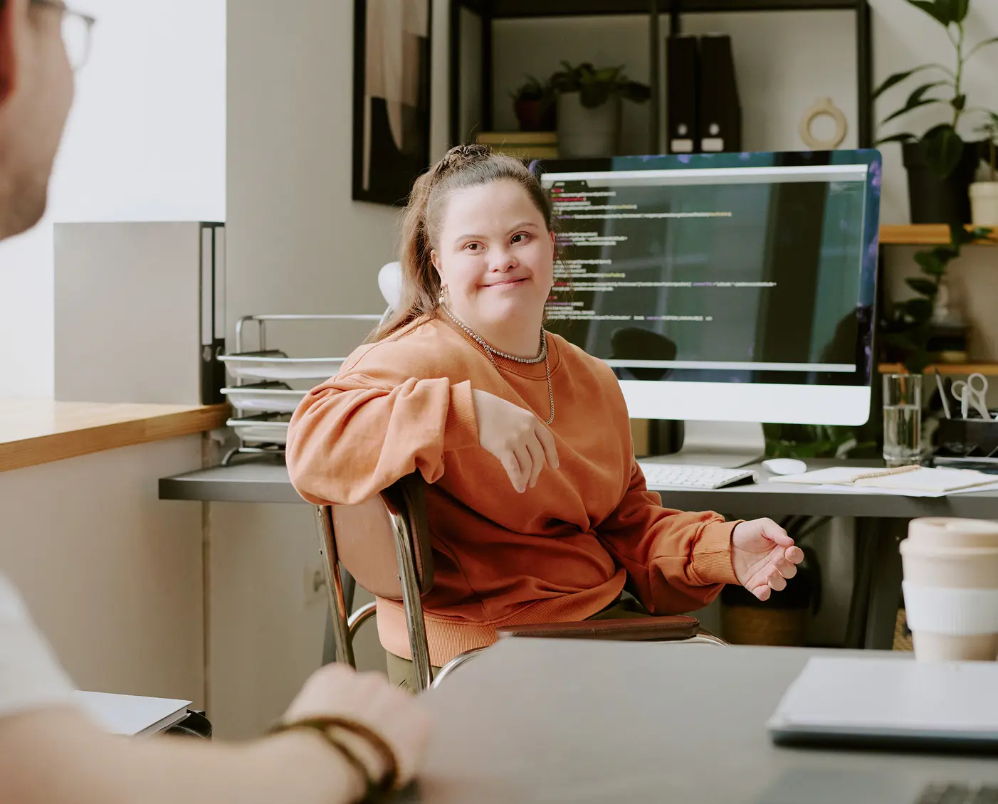 A woman (with Autism) in an orange sweatshirt sits near a computer monitor displaying lines of code. She smiles toward a coworker across the desk.