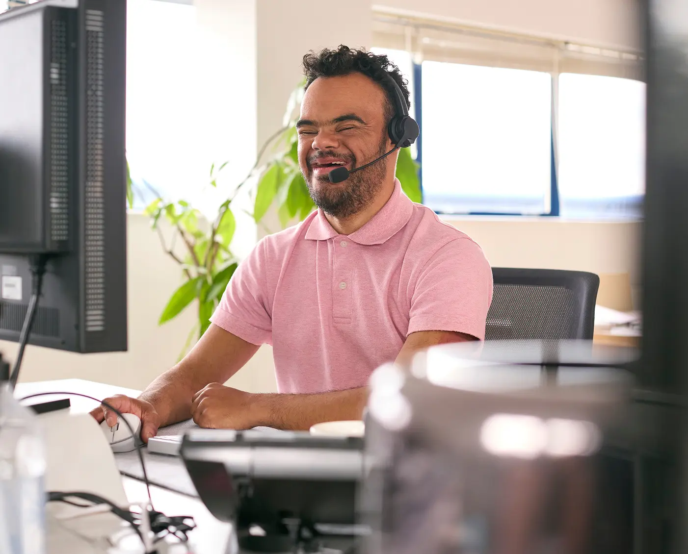 A blind man wearing a pink polo shirt and headset smiles while working on his computer in a bright room with sunlight coming through the window.